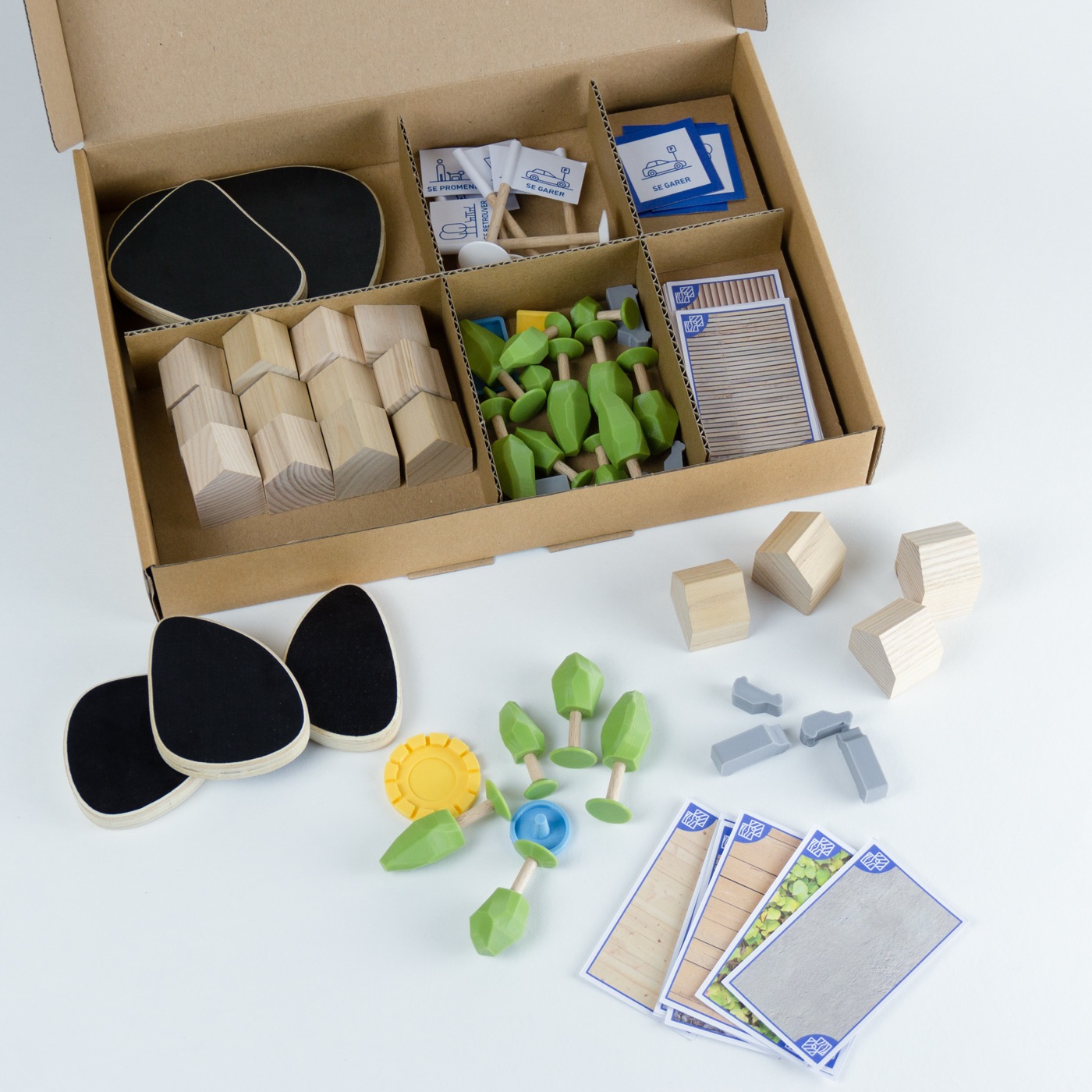 Une boîte de rangement compartimentée contenant les bâtiments en bois, les arbres, les véhicules, les drapeaux et des cartes d'échantillons de sols, une partie des pièces posées devant.