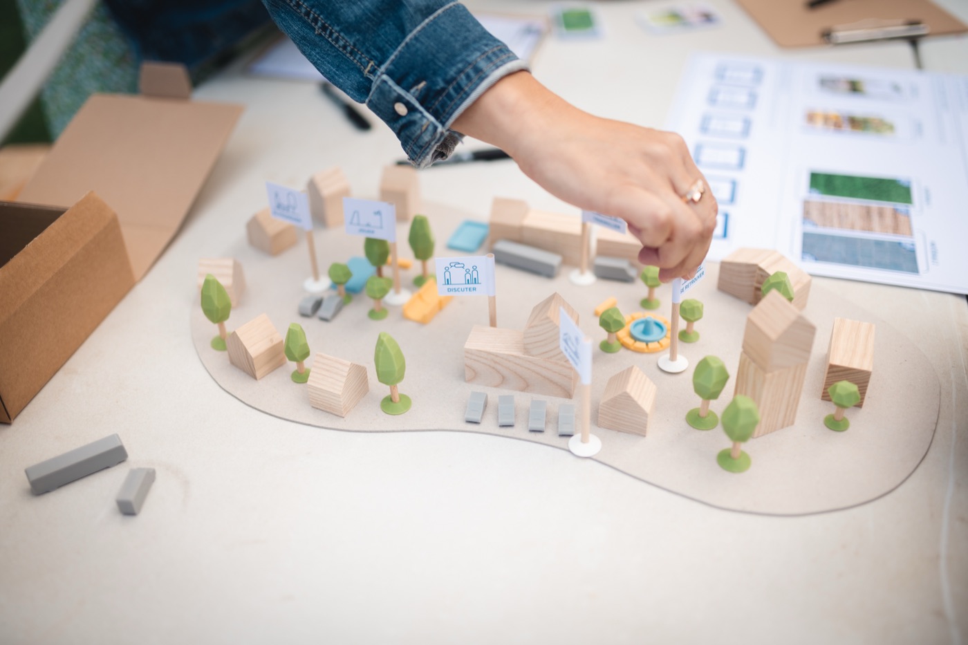 Une main plante un drapeau dans une maquette où voisinent des maisons en bois, des arbres, une file de véhicules gris et une fontaine posée sur l'agora.