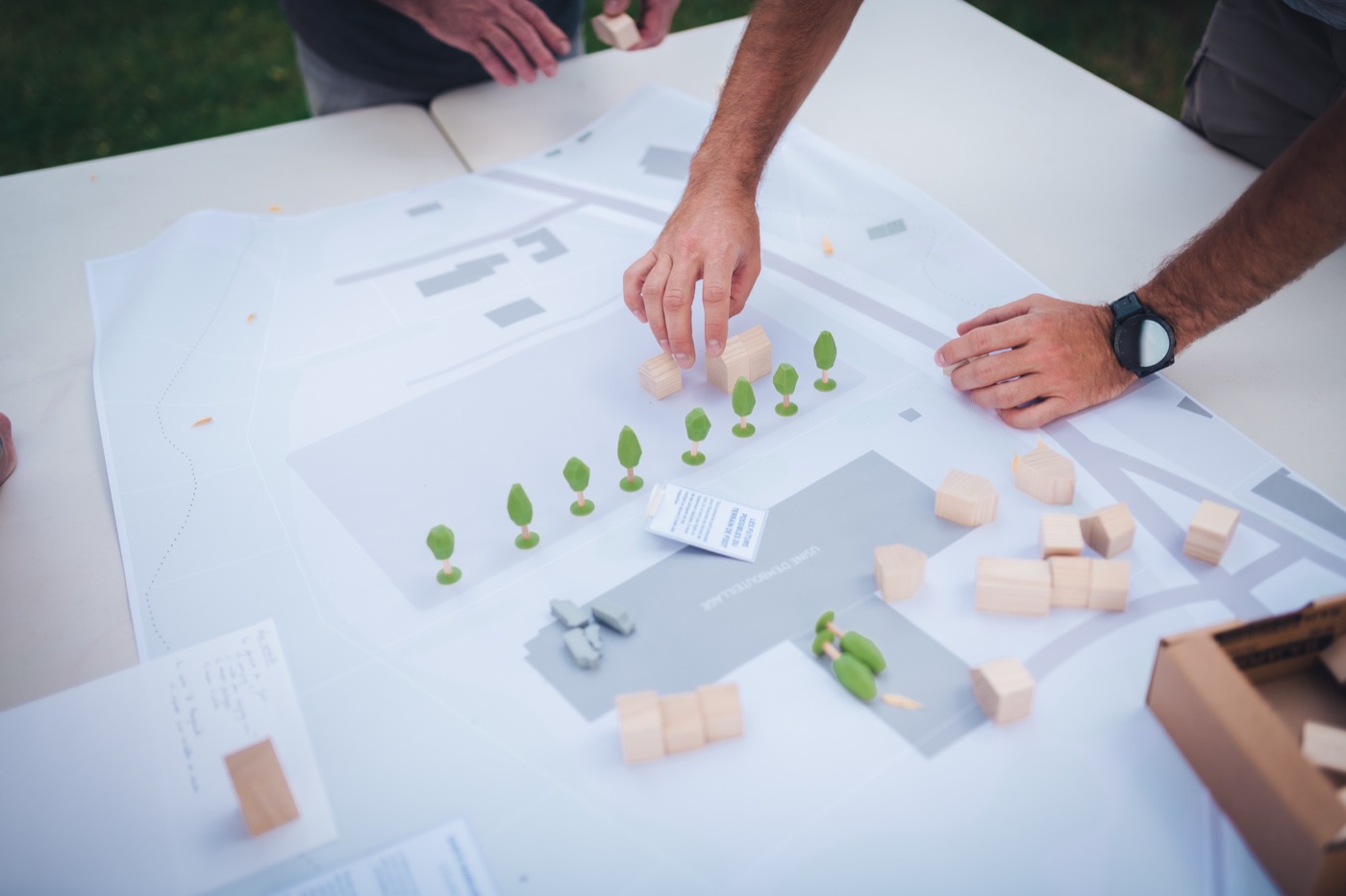 Deux personnes posent des volumes en bois et des arbres sur un grand plan de site imprimé, où une usine et des voies sont dessinées en gris.