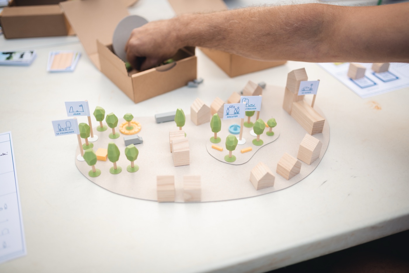 Une place composée sur un support ovale : arbres, agora jaune, fontaine bleue, bâtiments en bois, et de petits drapeaux nommant les usages, jouer, se promener, discuter, se garer.